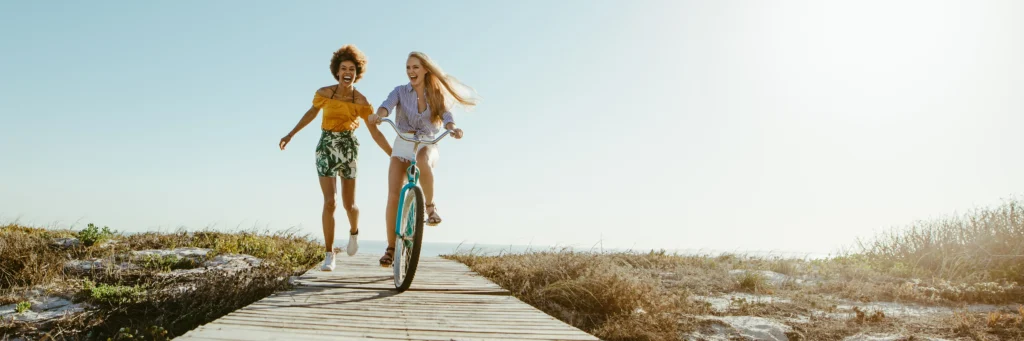 happy duo of friends riding a bike and jogging down a boardwalk from the beach